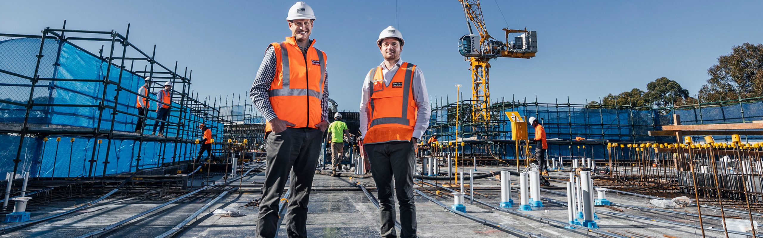 Directors Mathew and Daniel Edwards of Stephen Edwards Constructions, at a work site, in high viz, crane in background